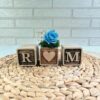 Tres cubos de madera personalizados con iniciales R-M y corazón central con rosa azul preservada, sobre superficie de mimbre natural