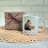 Taza blanca personalizada con foto de pareja y nombres Joana & Carlos, junto a paquete regalo con lazo granate sobre mantel de yute
