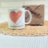 Taza blanca personalizada con mapa de amor en corazón, nombres y dedicatoria, sobre cesta de mimbre junto a regalo envuelto