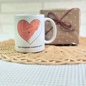 Taza blanca personalizada con mapa de amor en corazón, nombres y dedicatoria, sobre cesta de mimbre junto a regalo envuelto