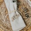 Marcasitios de madera personalizado con nombre Alba sobre servilleta beige y fondo de mimbre, detalle para boda
