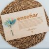 Placa de madera personalizada con dedicatoria para profesor grabada a láser, con texto enseñar y firma Laura sobre mantel de mimbre con flores secas azules