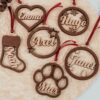 Bolas de Navidad personalizadas en madera de arce natural con nombres grabados a láser en diferentes formas: corazón, círculo con copos, estrella, bota y huella de mascota, con cintas rojas sobre fondo de mimbre