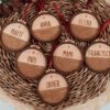 Mini bolas de Navidad personalizadas con nombres grabados en madera natural, con cinta roja, sobre cesta de mimbre con decoración navideña