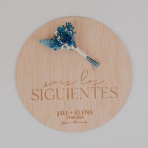 Placa redonda de madera personalizada con texto Sois los Siguientes, nombres Javi y Elena, fecha de boda grabados a láser y ramito de flores azules preservadas