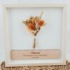 Cuadro de madera blanco con flores preservadas en tonos naranjas y naturales y placa grabada con mensaje personalizado para mamá, sobre fondo blanco